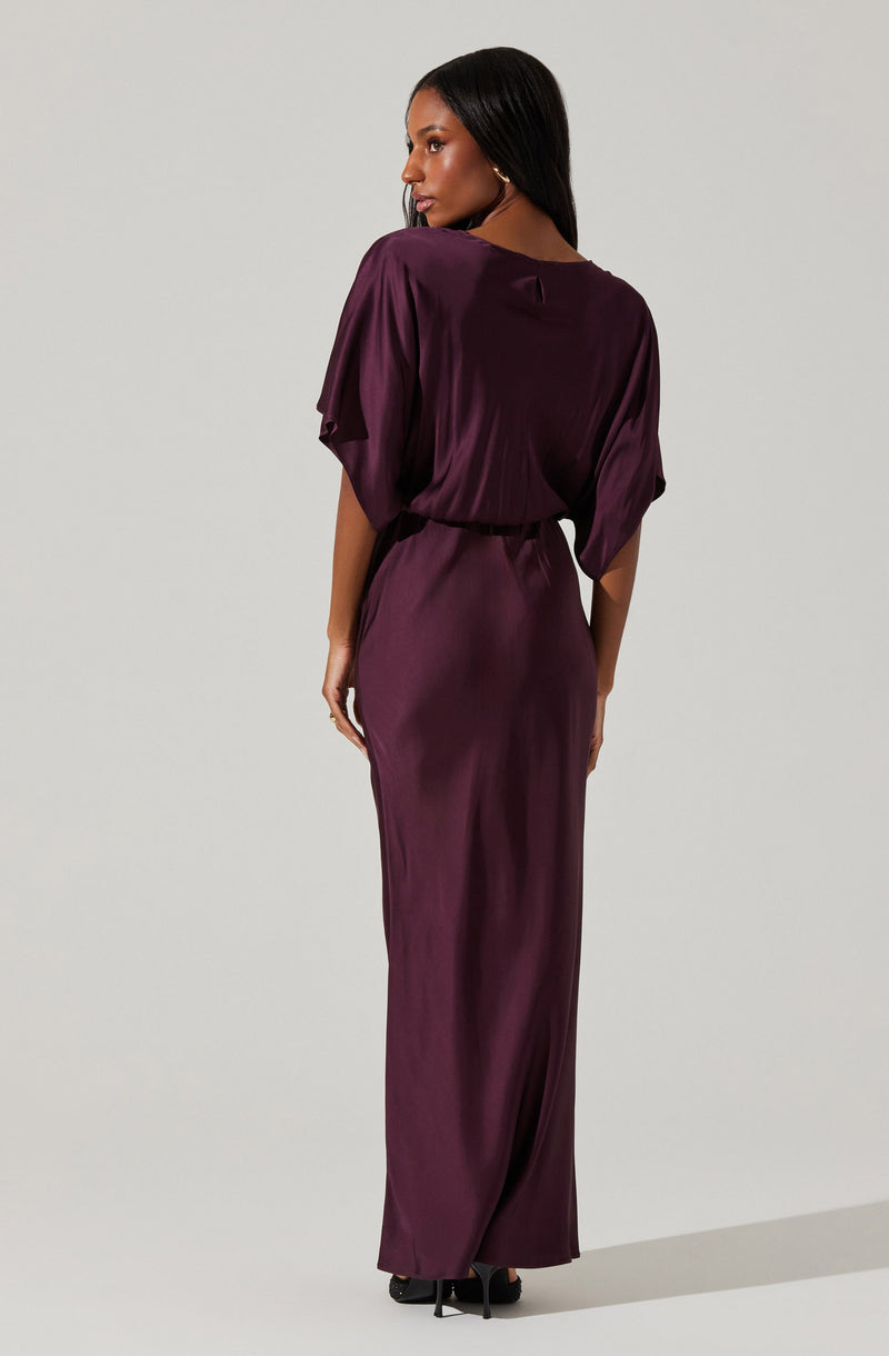 A woman is wearing a long, deep purple satin dress. The dress features a loose-fitting, short-sleeved top with a rounded neckline and a gathered waistline. The skirt is floor-length and flows down to the ground, creating an elegant silhouette. She is also wearing black high heels.