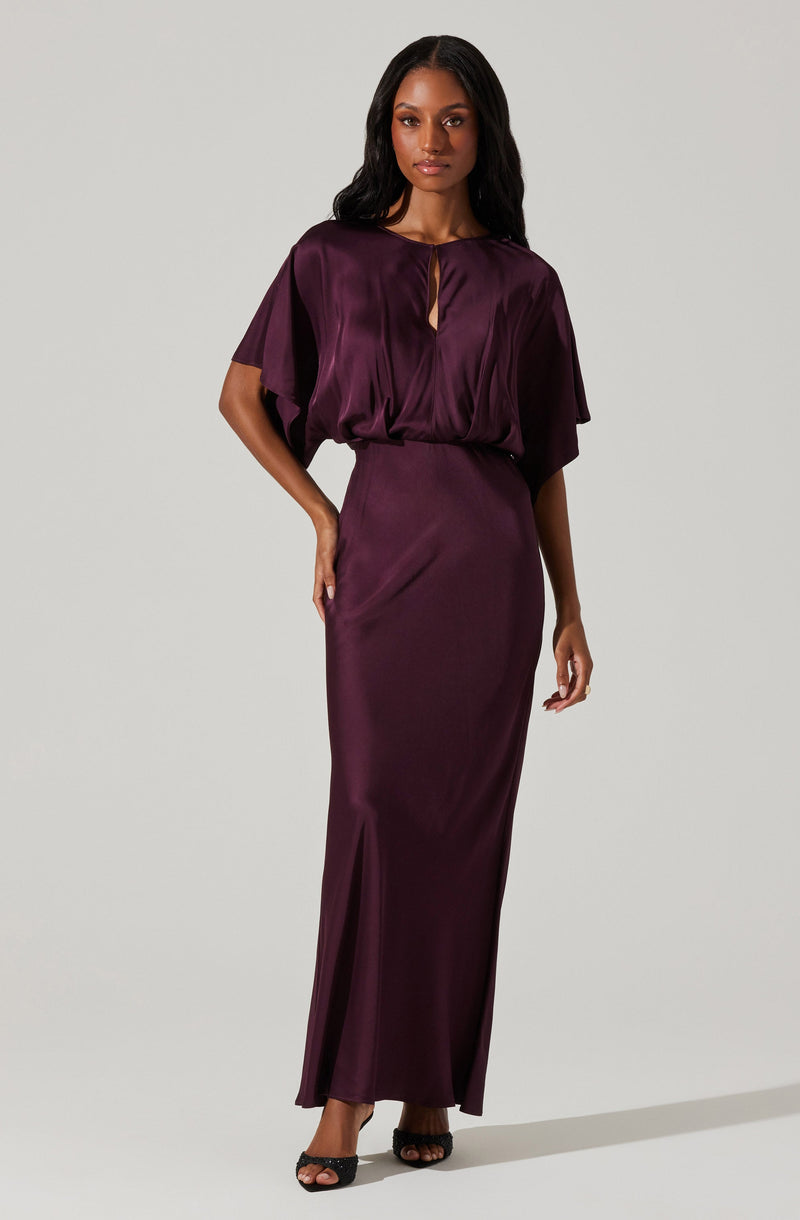 A woman is wearing a deep purple satin evening gown featuring a smooth, shiny fabric. The dress has a loose-fitting, draped bodice with a keyhole neckline and short, wide sleeves. The skirt is fitted at the waist and extends to the floor with a subtle side slit.