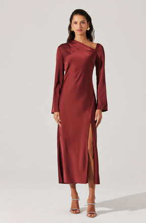 A woman is wearing a long, elegant gown made of shiny satin fabric in a deep burgundy color. The dress features long sleeves, an asymmetrical neckline, and a high slit on one side that reveals her leg. She is also wearing strappy high-heeled sandals.