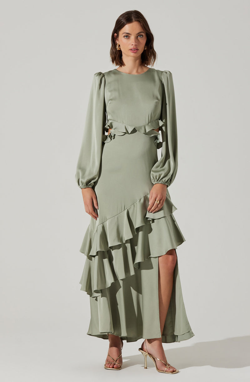 A woman is wearing a long, pale green dress with puffed long sleeves that are gathered at the wrists. The dress features cut-out details on the sides of the waist with ruffled accents, and a layered, ruffled hemline that cascades asymmetrically down the front and back, revealing a high slit on one side. She is also wearing gold high-heeled sandals and gold hoop earrings.