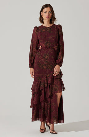 A woman is wearing a long, burgundy dress with an intricate floral pattern in a darker shade. The dress features puffed long sleeves with elastic cuffs, a ruffled front detail at the waist, and a high slit on one side revealing her leg. She is also wearing black strappy high-heeled sandals.