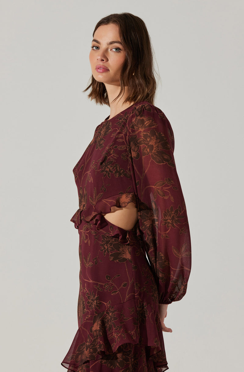 A woman is wearing a long-sleeved, burgundy dress with a floral pattern in shades of brown and orange. The dress features gathered fabric at the cuffs and a ruffled hem that adds a decorative touch.