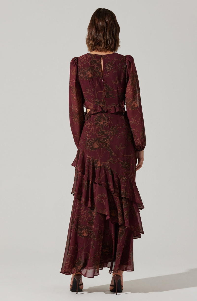 A woman is wearing a long, flowing dress with a deep burgundy color and an intricate floral pattern in shades of gold and brown. The dress features long, puffed sleeves and a fitted waist, with layered ruffles cascading down the length of the skirt.
