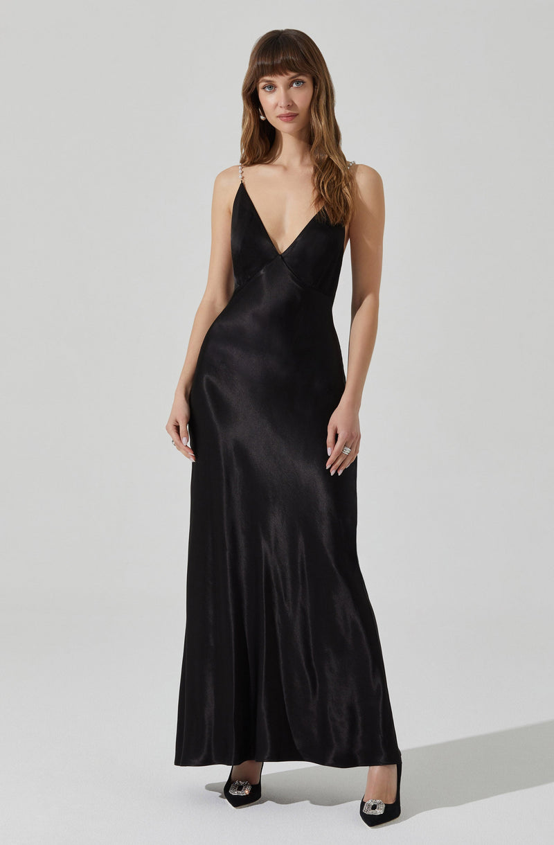 A woman is wearing a long, black satin gown with a deep V neckline. The dress features thin, beaded shoulder straps. She is also wearing black high-heeled shoes with embellishments on the toes.