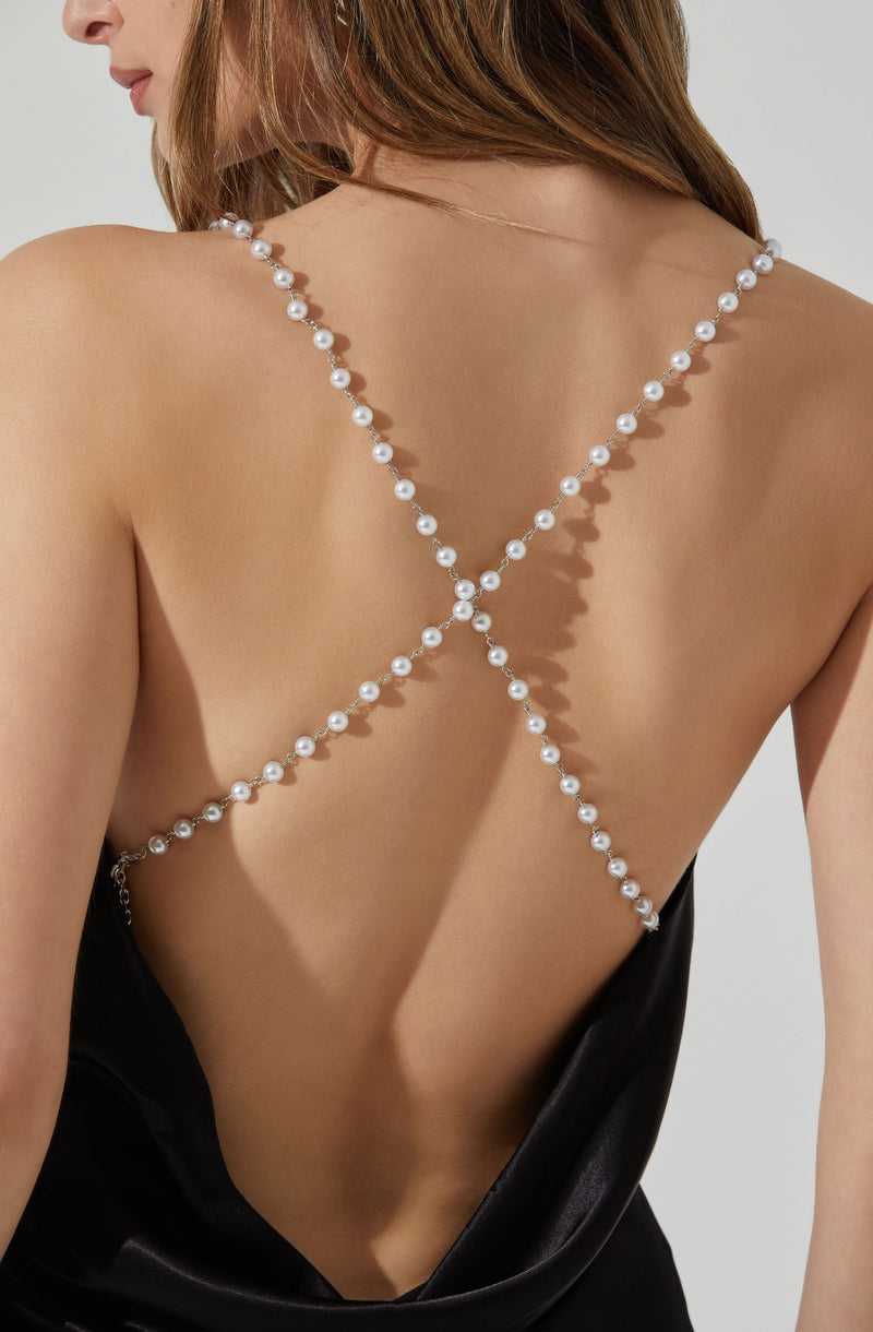 A woman is wearing a black sleeveless outfit with an open back, decorated with a delicate strappy necklace made of white pearls. The necklace crosses at the back, creating an X shape.