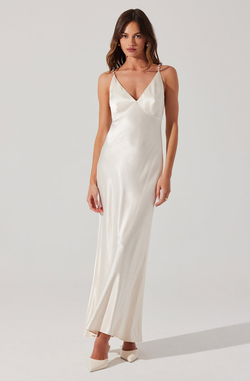 A woman is wearing a long, elegant satin dress in an ivory or champagne color. The dress features a V-neckline, thin spaghetti straps, and a fitted silhouette that flows down to the floor. She is also wearing matching pointed-toe heels.