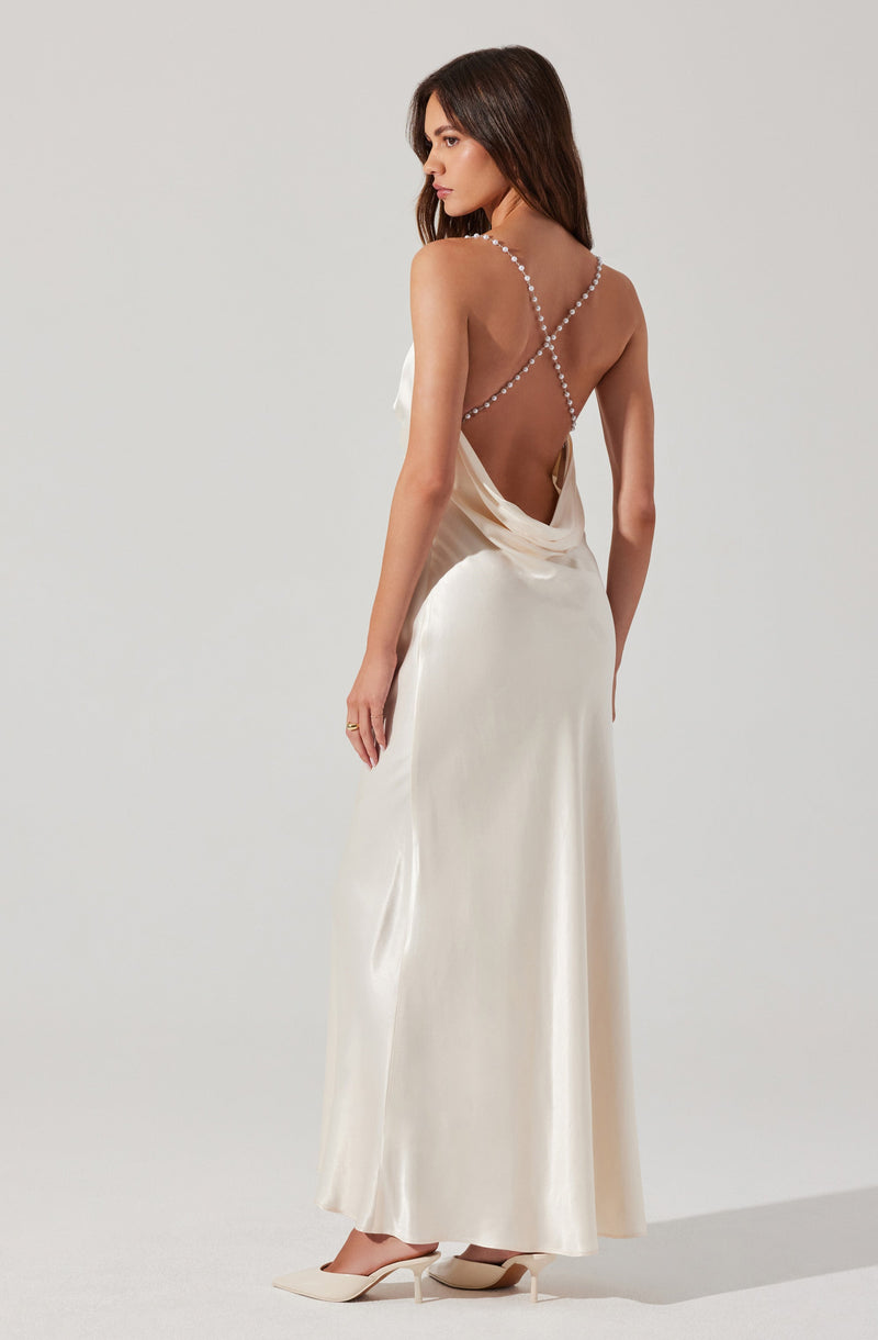 A woman is wearing a long, elegant cream-colored satin dress with a backless design. The dress features thin, beaded straps that cross over her back, creating a delicate and sophisticated look. She is also wearing matching pointed-toe heels.