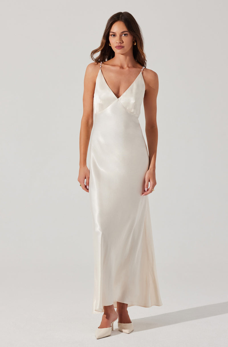 A woman is wearing a long, elegant, ivory satin gown with a deep V-neckline. The dress features thin, beaded spaghetti straps and a fitted silhouette that flows down to the floor. She is also wearing matching ivory shoes with a pointed toe.