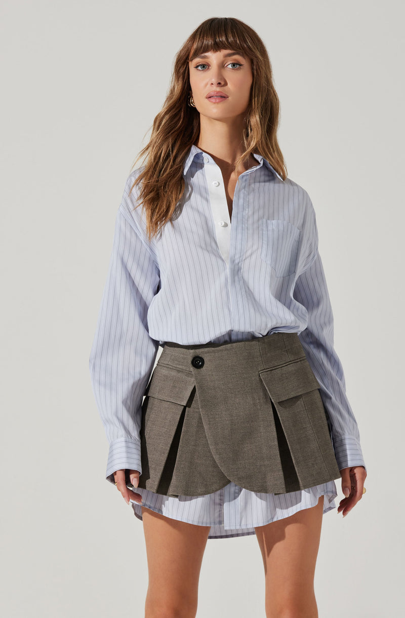 A woman is wearing a light blue, long-sleeved, button-up shirt with thin white vertical stripes. The shirt has a collar and a chest pocket on the left side. She is also wearing a high-waisted, grayish-brown plaid mini skirt with large front pockets that have flaps, and decorative pleats in the front.