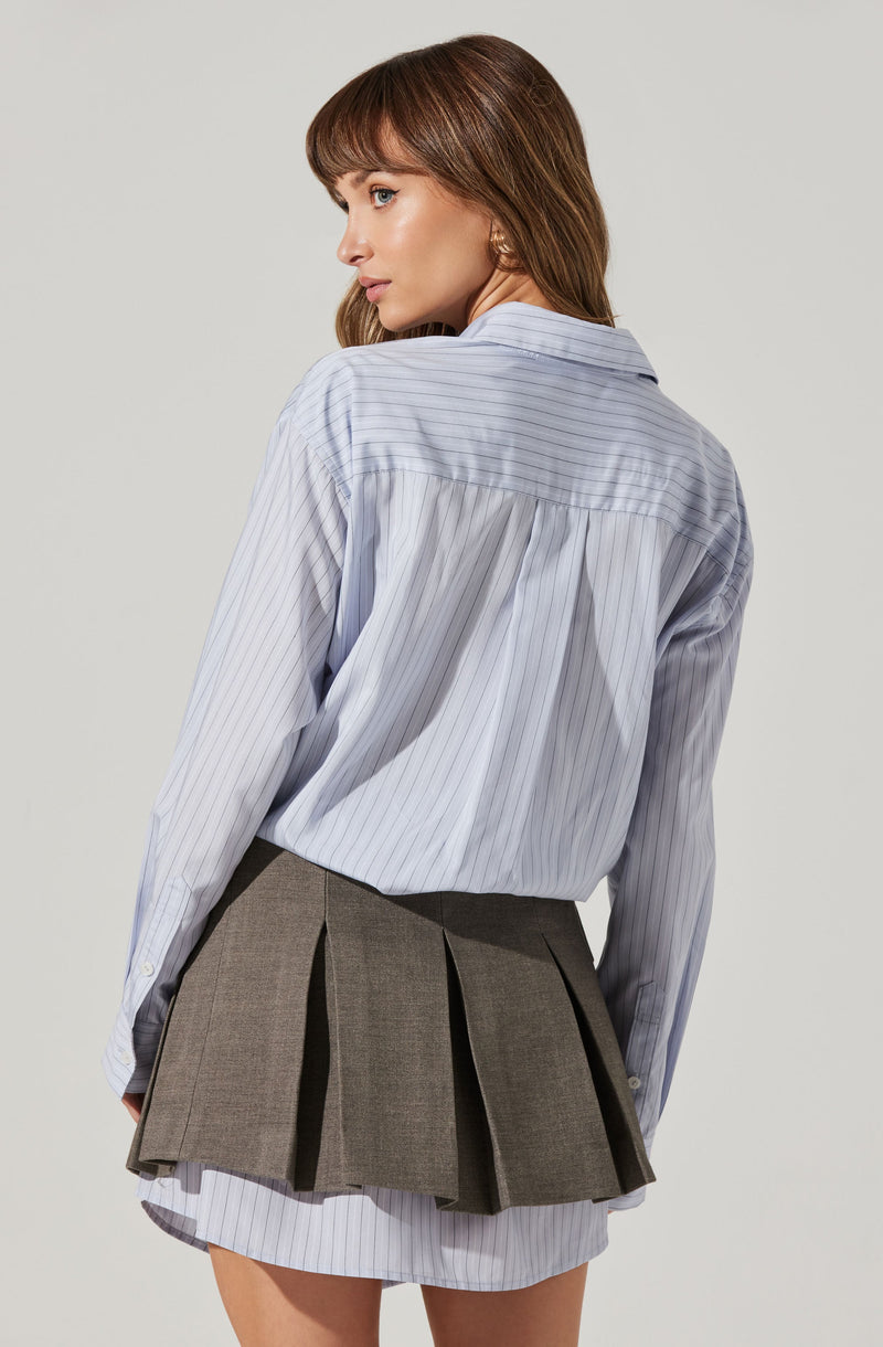 A woman is wearing a light blue, long-sleeved, collared shirt with thin vertical white stripes. The shirt is slightly oversized and tucked into a layered, pleated gray skirt. The skirt is short, with a portion of a matching striped fabric visible at the hem underneath the pleated layer.