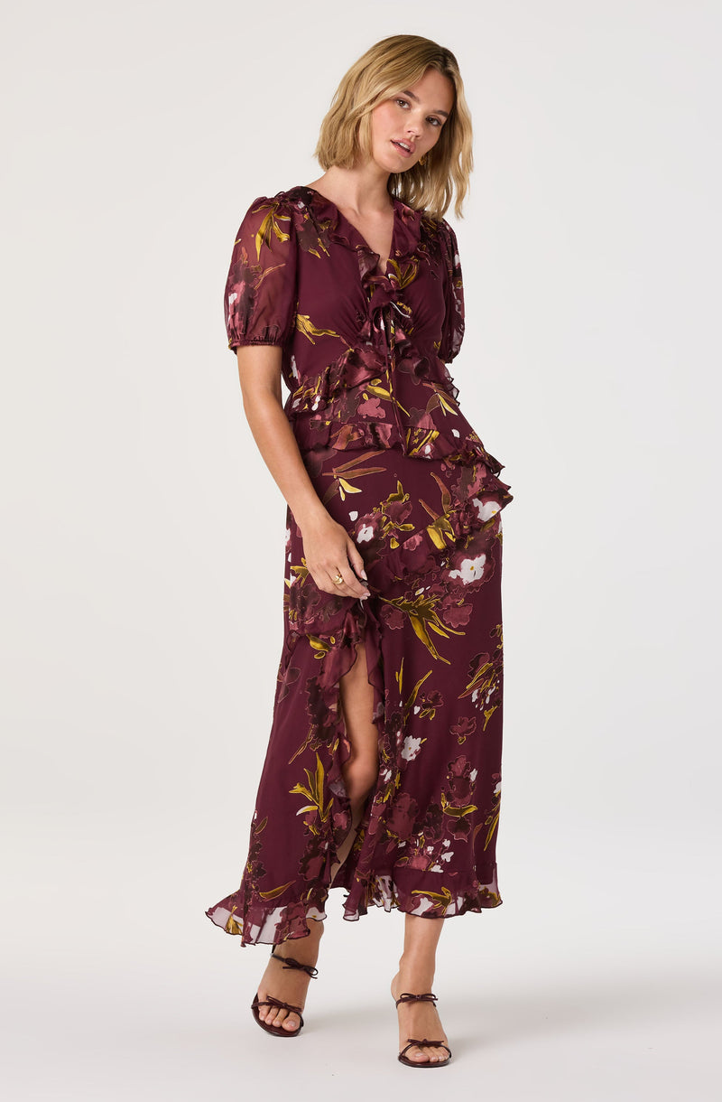 A woman is wearing a matching two-piece outfit with a deep burgundy background and a floral pattern featuring white, pink, and yellow flowers with green leaves. The top has short puffed sleeves, a V-neckline with ruffles, and a tie detail at the front. The skirt is long and flowing with a slit up one side, revealing part of her leg. The fabric appears lightweight and sheer. She is also wearing strappy black high-heeled sandals.