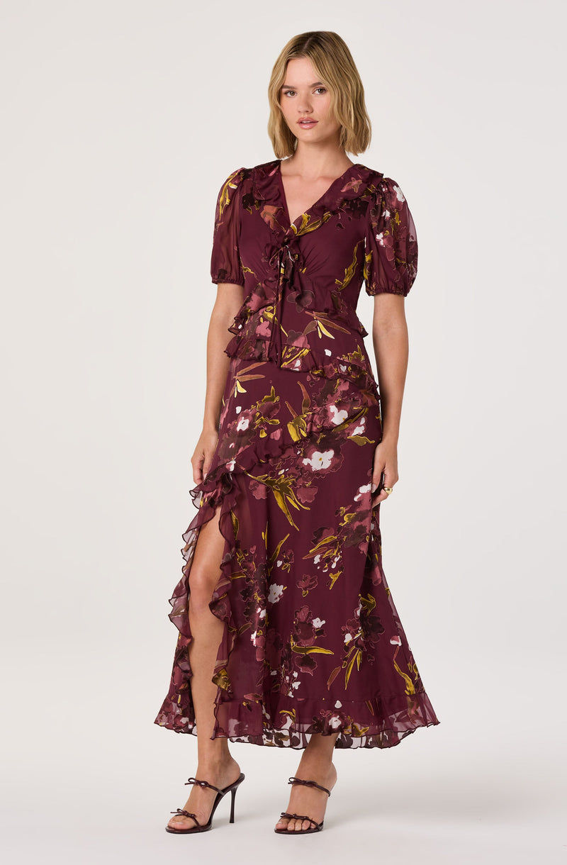 A woman is wearing a long, flowing dress with a floral pattern. The dress features a deep burgundy color with yellow and white flowers and green leaves. It has short, puffed sleeves and a V-neck with a bow tie at the front. The skirt has ruffled details along the side slit, which reveals part of her leg. She is wearing black high-heeled sandals.