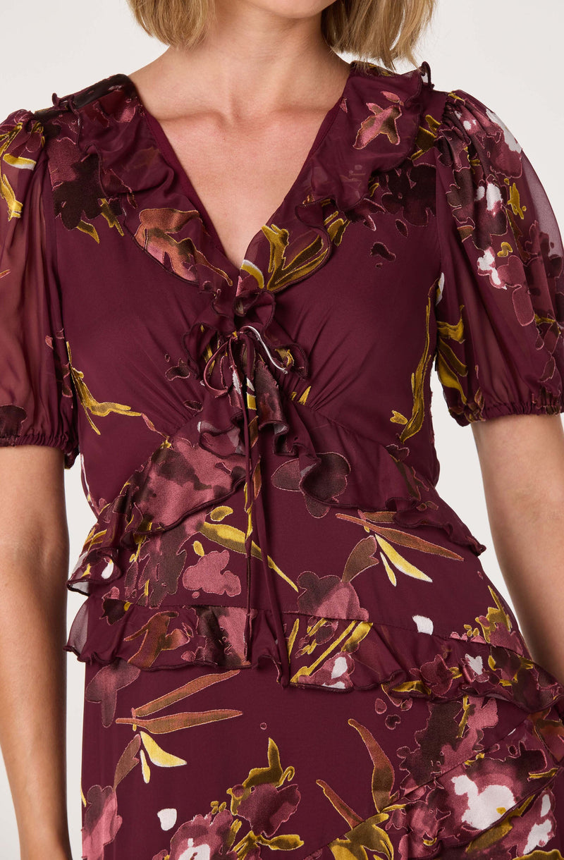 A woman is wearing a burgundy dress with a floral pattern in shades of pink, white, and gold. The dress features puffed short sleeves, a ruffled V-neckline, and ruffle details across the bust and waist. It is made of a shiny, silky fabric that drapes smoothly and has a tied bow at the center of the neckline.