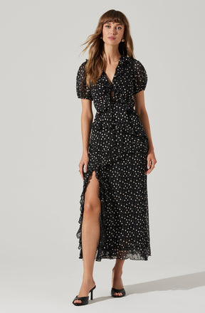 A woman is wearing a black dress with a white polka dot pattern. The dress features a V-neckline, puffed short sleeves, and a tiered ruffle detail that runs down the front and around the hem. It has a high slit on one side, revealing her leg. She is also wearing black high-heeled sandals.