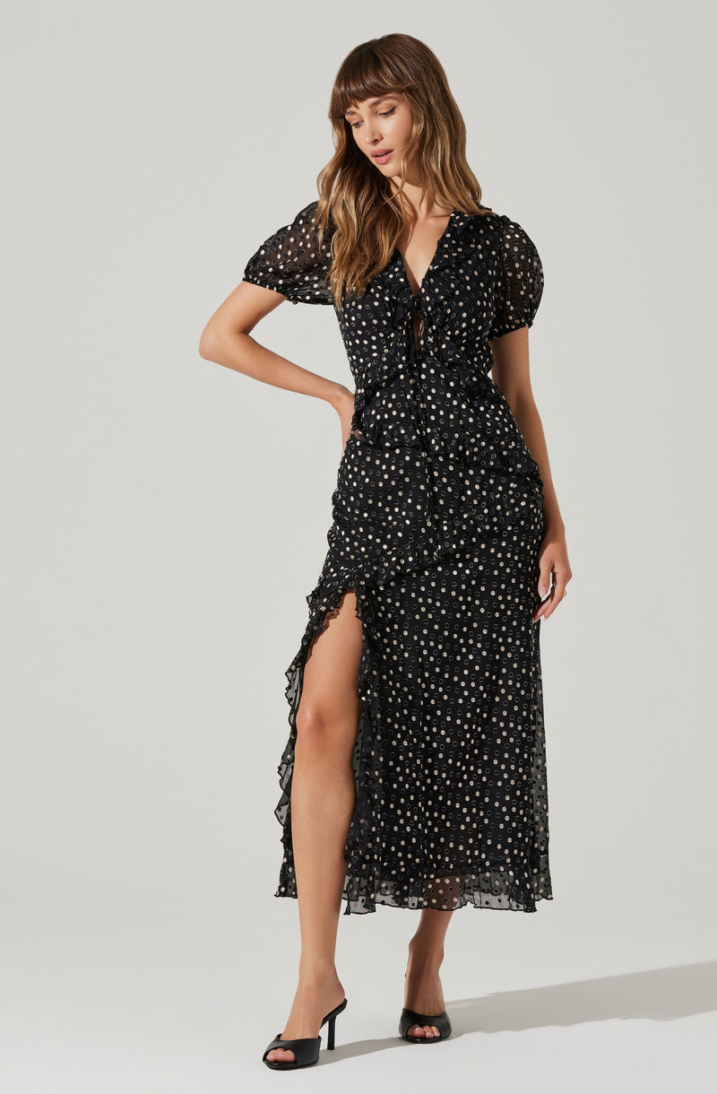 A woman is wearing a black, midi-length dress with a sheer, dotted pattern. The dress features a ruffled front with a tie at the neckline, short puffy sleeves, a high slit on the left side revealing her leg, and a ruffled hem. She is also wearing black high-heeled shoes.