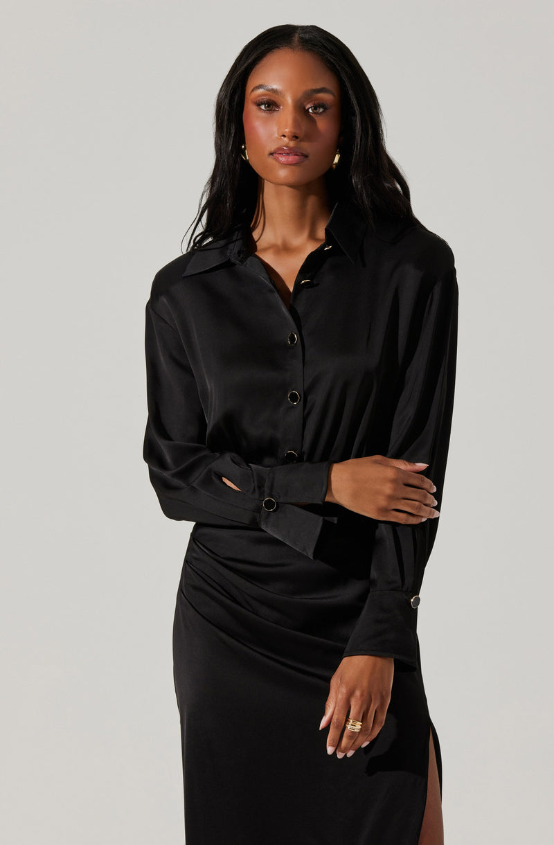 A woman is wearing a long-sleeved black silk blouse with a classic collar and buttons down the front. The blouse has slightly puffed sleeves with buttoned cuffs. She is also wearing a black skirt with a side slit.