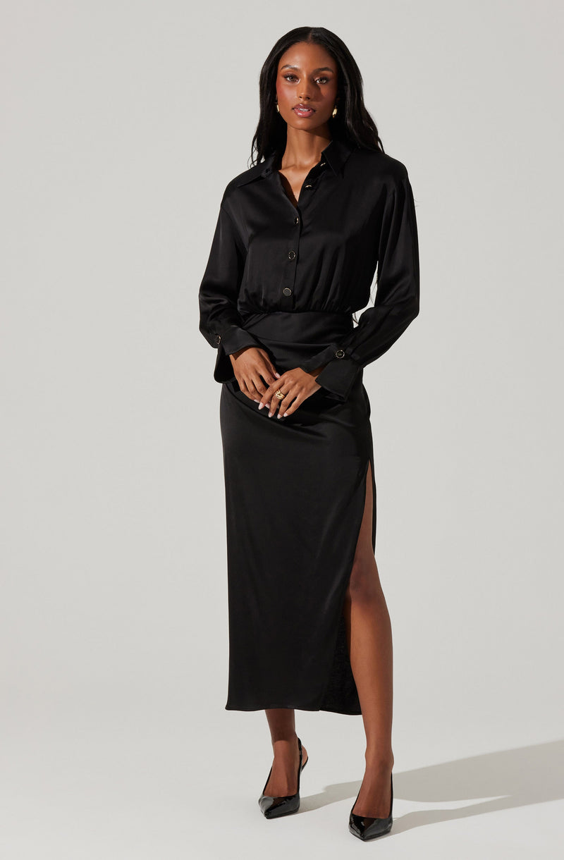 A woman is wearing a long-sleeved black satin button-up shirt with a collar, paired with a matching black satin skirt that has a high slit on the left side, exposing her leg. She is also wearing black pointed-toe high heels.