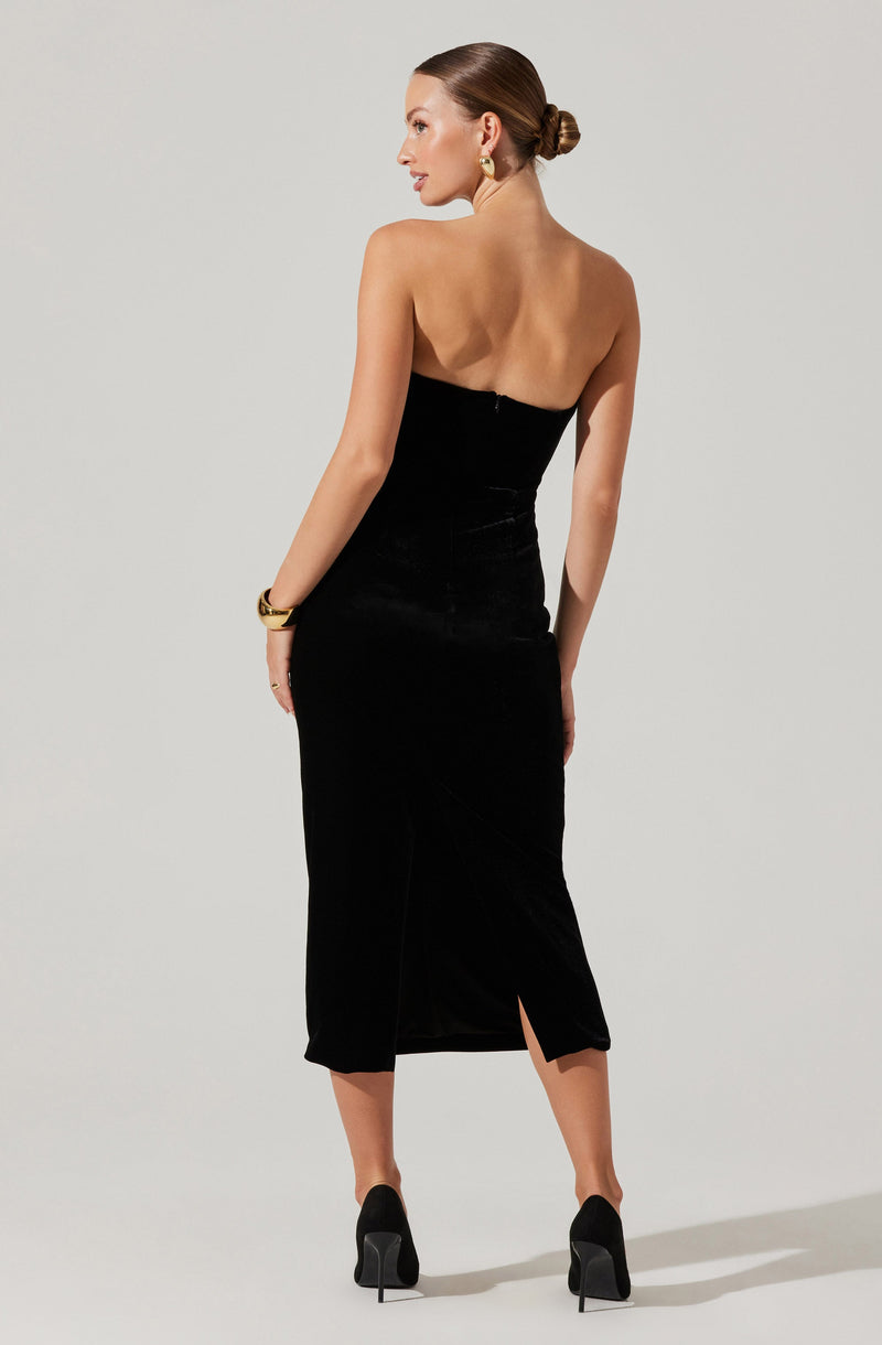 A woman is wearing a strapless black dress that is fitted at the waist and extends to a mid-calf length with a small slit at the back. She has accessorized with gold earrings and a bracelet, and is wearing black high heels. Her hair is styled in a sleek, low bun.