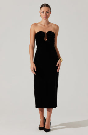 A woman is wearing a strapless, black, floor-length gown with a keyhole cutout in the center of the bodice. The top of the gown features gathered fabric around the bust area. She accessorizes with gold earrings, a gold bracelet, and rings. She is wearing black high heels.