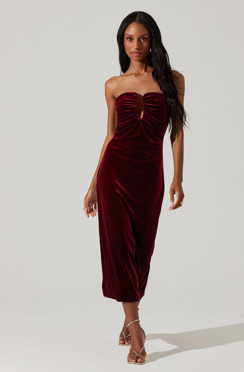A woman is wearing a strapless, deep red velvet dress that features a ruched bodice with a keyhole opening in the center. The dress is midi-length, fitted at the waist, and flows down to her ankles. She is also wearing open-toed, high-heeled sandals with thin straps.