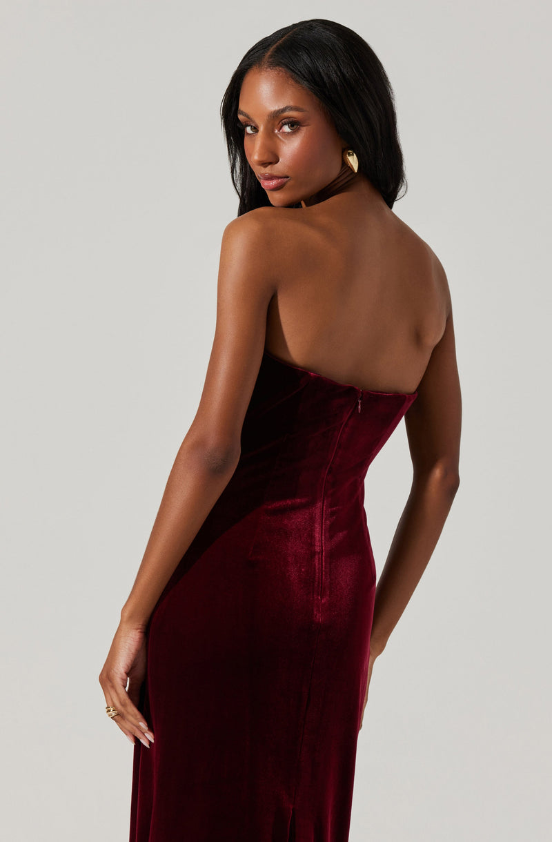 A woman is wearing a strapless, deep red velvet gown that fits closely to her body. The dress has a smooth, shiny texture and a zipper closure at the back. She accessorizes with gold earrings and has dark, straight hair.