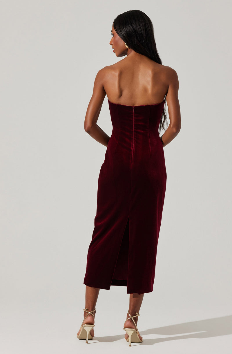 A woman is wearing a strapless, deep red velvet dress that is fitted at the waist and extends to mid-calf length. The dress features vertical seam details and a back slit at the hem. She pairs the outfit with beige strappy high-heeled sandals.