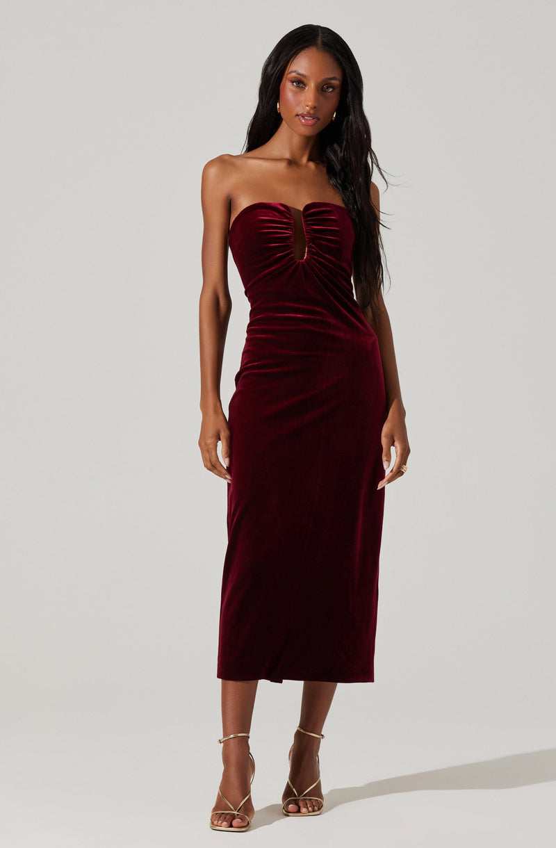 A woman with long dark hair is wearing a strapless, deep red velvet midi dress with a gathered, ruched bodice and a keyhole cutout at the center. She is also wearing gold strappy high-heeled sandals. The background is plain and light-colored.