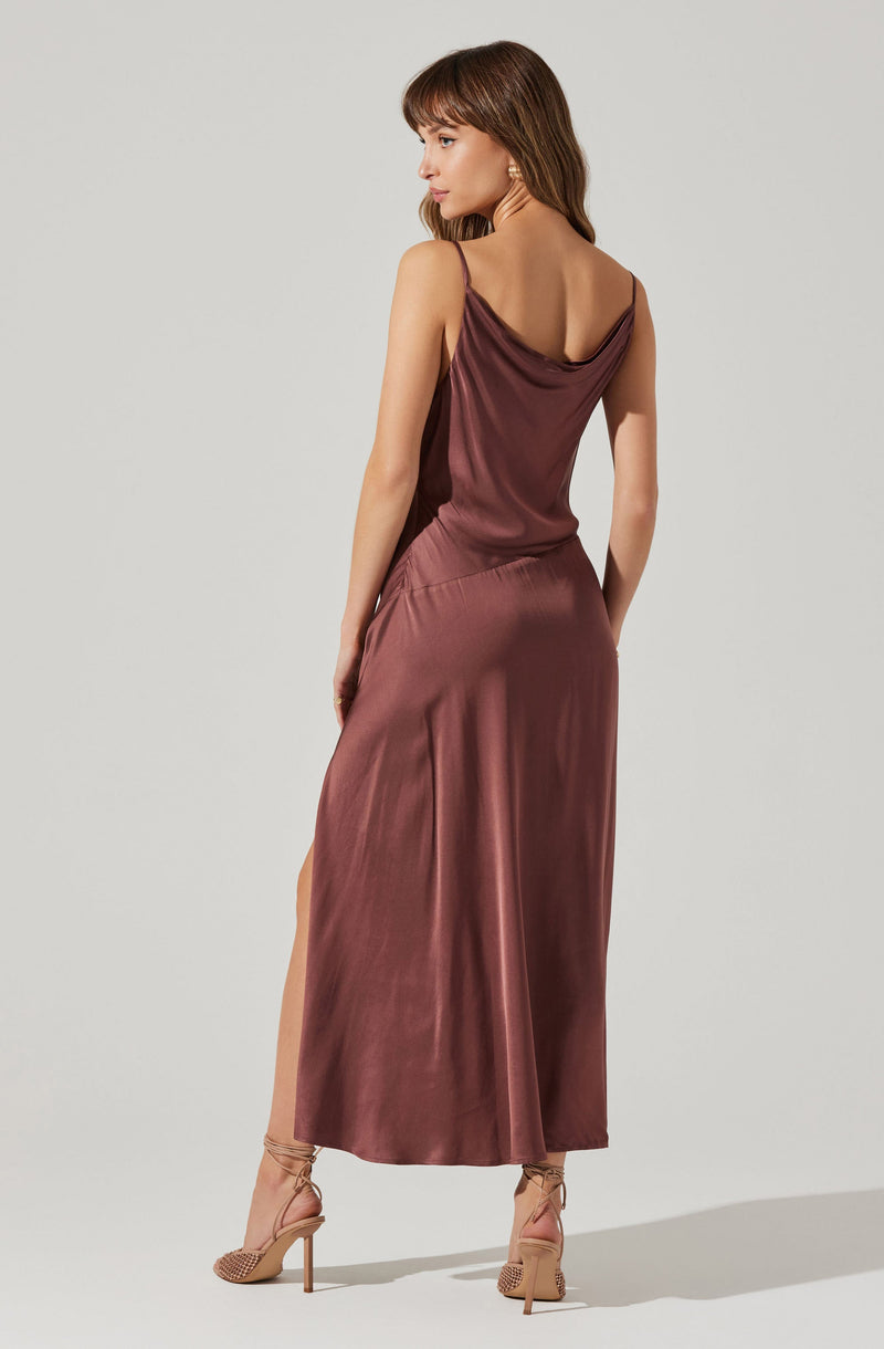 A woman is wearing a sleeveless, silky, mauve-colored dress with thin spaghetti straps and a cowl neckline. The dress has a flowy silhouette and a high slit on one side, revealing her leg. She is also wearing nude, lace-up high-heeled shoes.