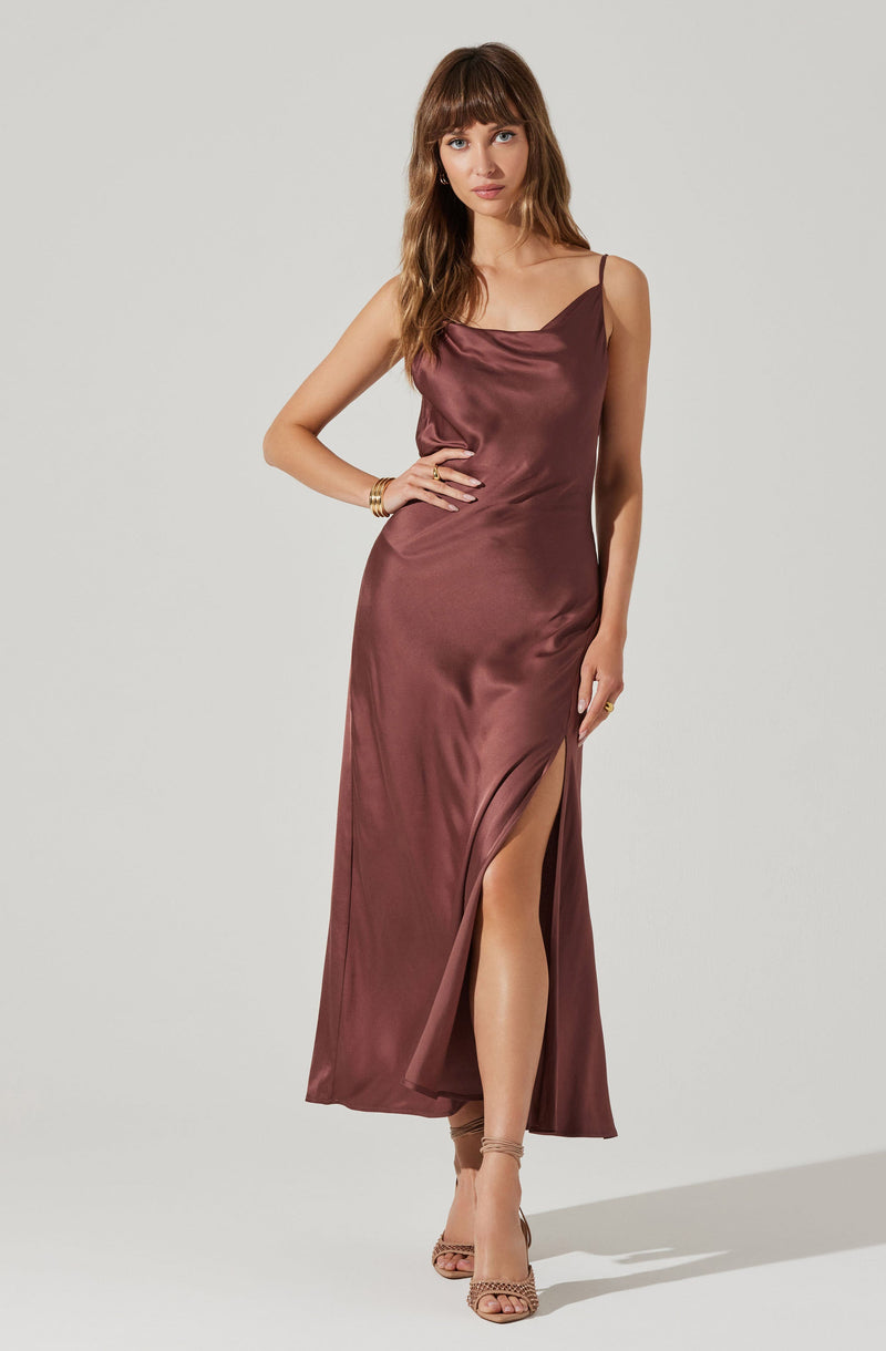 A woman is wearing a full-length satin dress in a deep mauve or wine color with thin spaghetti straps. The dress features a draped cowl neckline and a high slit on the left side, revealing her leg. She is accessorized with a gold bracelet and a ring, and is wearing nude high-heeled sandals.