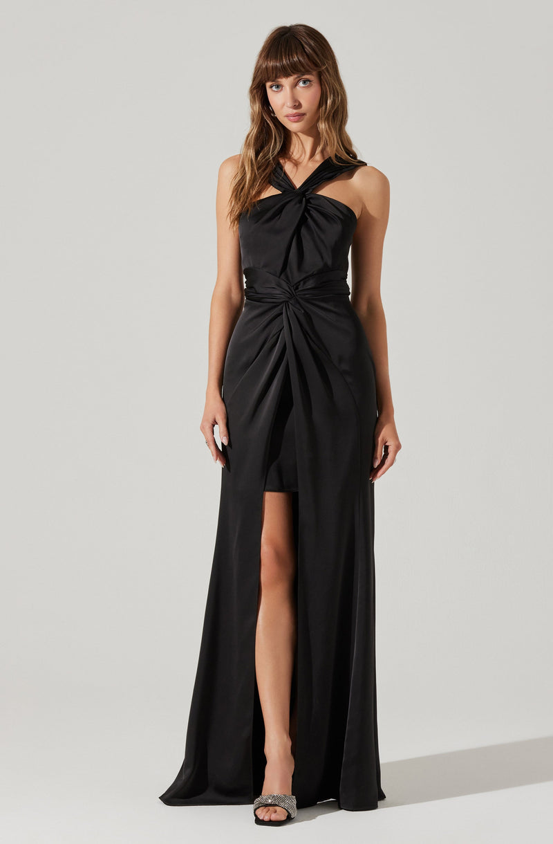 A woman is wearing a sleeveless, floor-length black gown with a high slit. The dress features a twisted knot design at the waist and a gathered detail at the bust with crossover straps at the shoulders. She is also wearing open-toe high heels with embellishments.