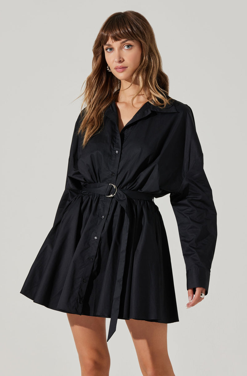 A woman is wearing a black, long-sleeved shirt dress made from a shiny, satin-like fabric. The dress features a pointed collar, button-down front, and a matching belt with a circular metal buckle at the waist. The skirt portion of the dress is gathered and flares out slightly, ending above the knees.