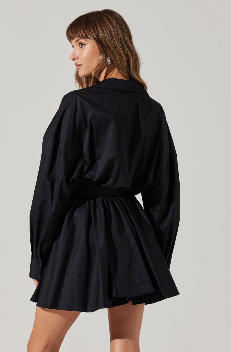 A woman is wearing a black shirt with long, loose sleeves and a collar. The shirt has a relaxed fit and is tucked into a matching black, high-waisted skirt that is pleated and falls above the knees.