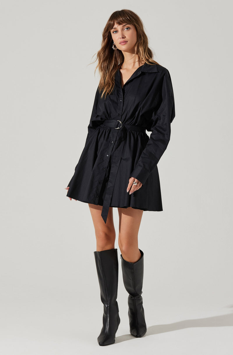 A woman is wearing a black long-sleeved shirt dress with a collar and button-down front. The dress has a matching fabric belt with a silver buckle tied around her waist. She pairs the dress with knee-high black boots.