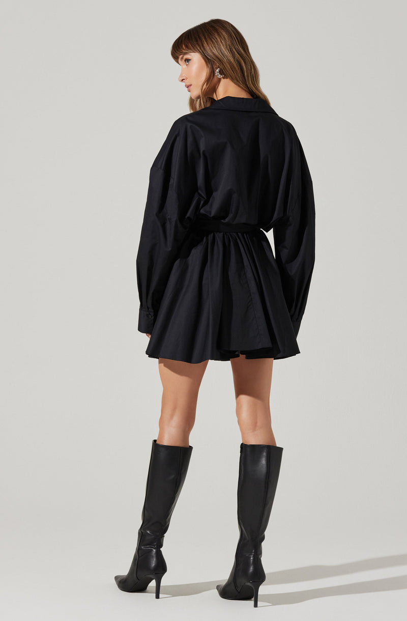 A woman is wearing a loose-fitting, long-sleeved black shirt with a collar, paired with a high-waisted black skirt that has a flared, pleated design. She is also wearing tall black heeled boots that reach just below the knee.