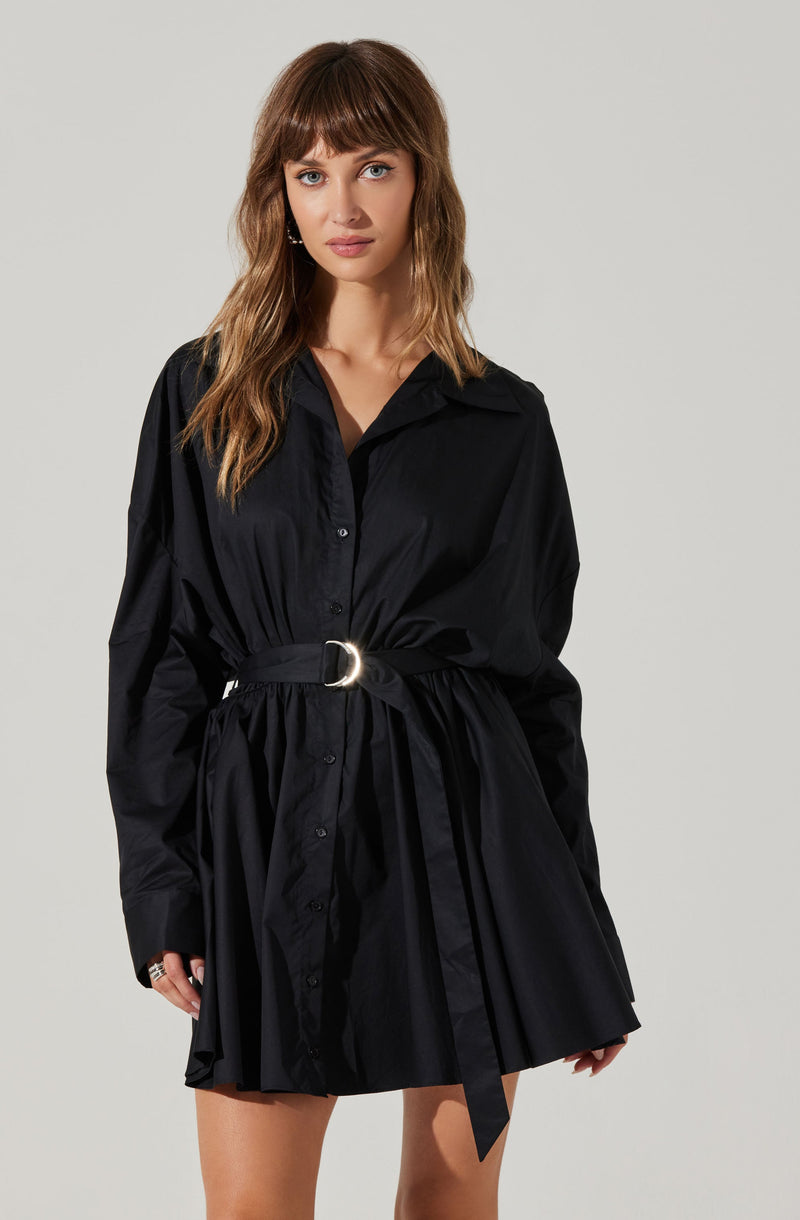 A woman is wearing a black shirt dress with a collared neckline, long sleeves, and button-down front. The dress features a matching fabric belt with a silver D-ring buckle, cinching the waist and creating a gathered, pleated skirt.