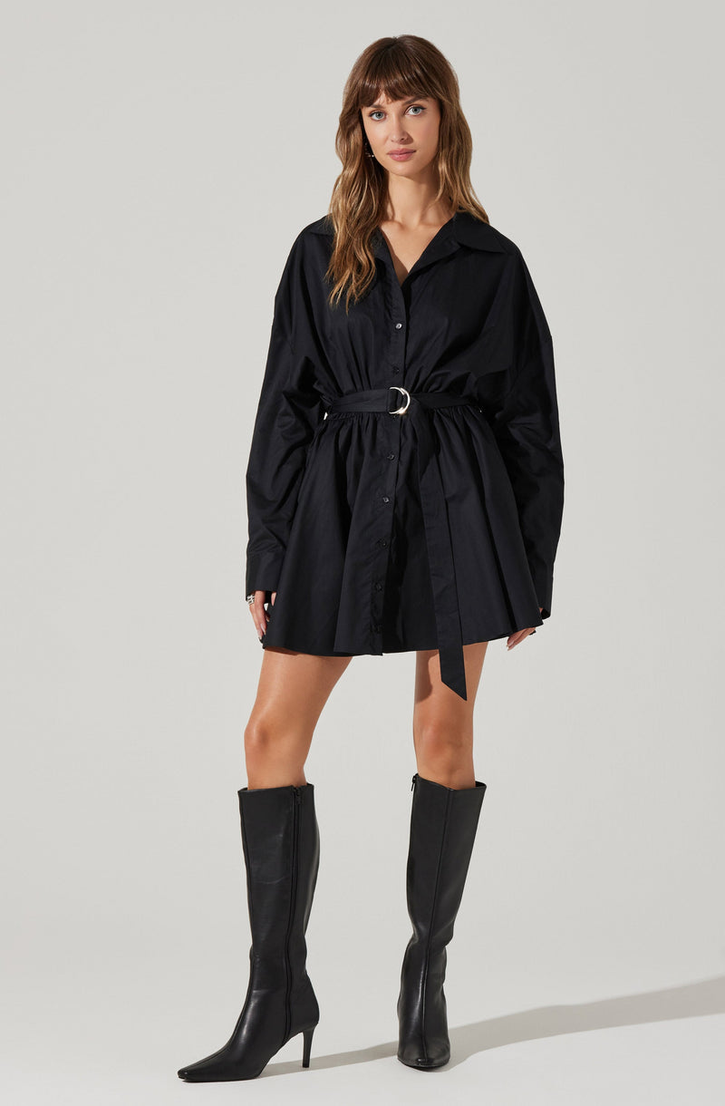 A woman is wearing a black, long-sleeved shirt dress with a collar and buttons down the front. The dress features a gathered waist with a matching fabric belt that has a silver buckle. She is also wearing tall black leather boots that reach just below the knees and have pointed toes and high heels.