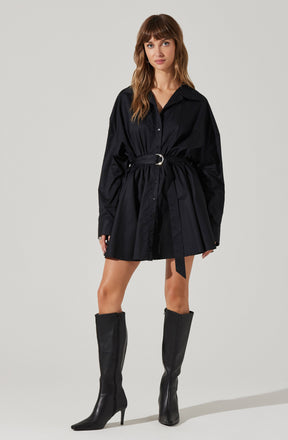 A woman is wearing a black, long-sleeved shirt dress with a collar and buttons down the front. The dress features a gathered waist with a matching fabric belt that has a silver buckle. She is also wearing tall black leather boots that reach just below the knees and have pointed toes and high heels.