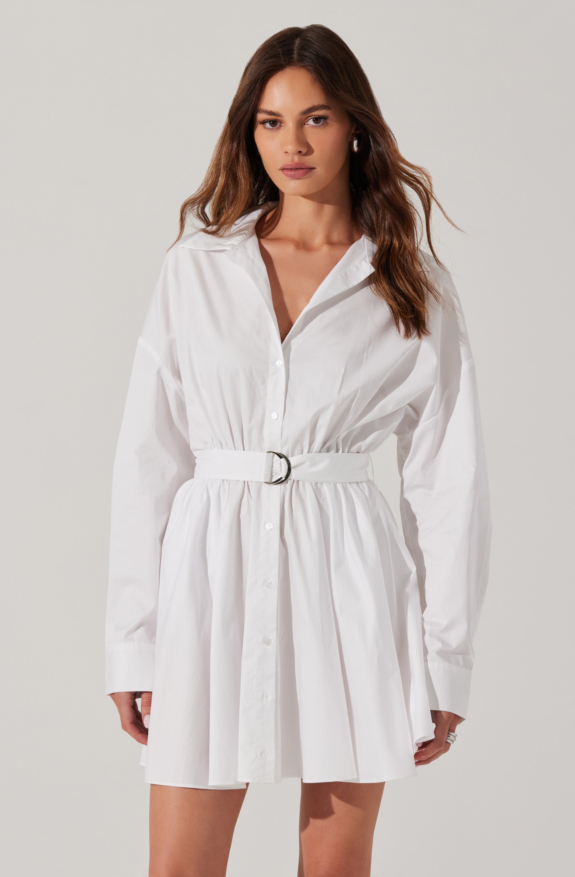 A woman is wearing a white, long-sleeved shirt dress with a collar and button-down front. The dress features a gathered waist with a matching fabric belt that has a silver buckle. The skirt portion of the dress is pleated, creating a slightly flared silhouette. The woman's hair is long and wavy, and she has a neutral expression on her face.