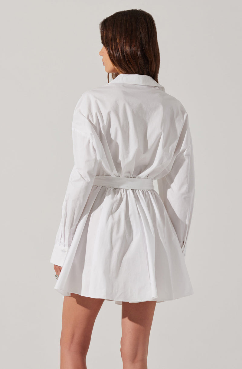 A woman is wearing an oversized white shirt dress with long sleeves, a collar, and a wide waistband that cinches at the waist. The dress features a gathered skirt that falls above the knees.