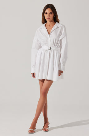 A woman is wearing a white long-sleeved shirt dress with a collar and a front zipper. The dress has a gathered waist with a matching belt featuring a circular buckle and a pleated skirt. She is also wearing beige high-heeled sandals.