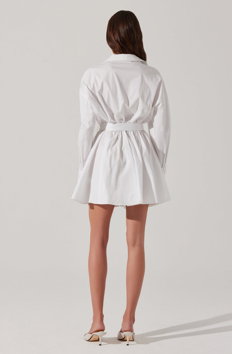 A woman is wearing a white long-sleeve blouse dress with a cinched waist and a gathered, flared skirt. She is also wearing white high-heeled shoes.