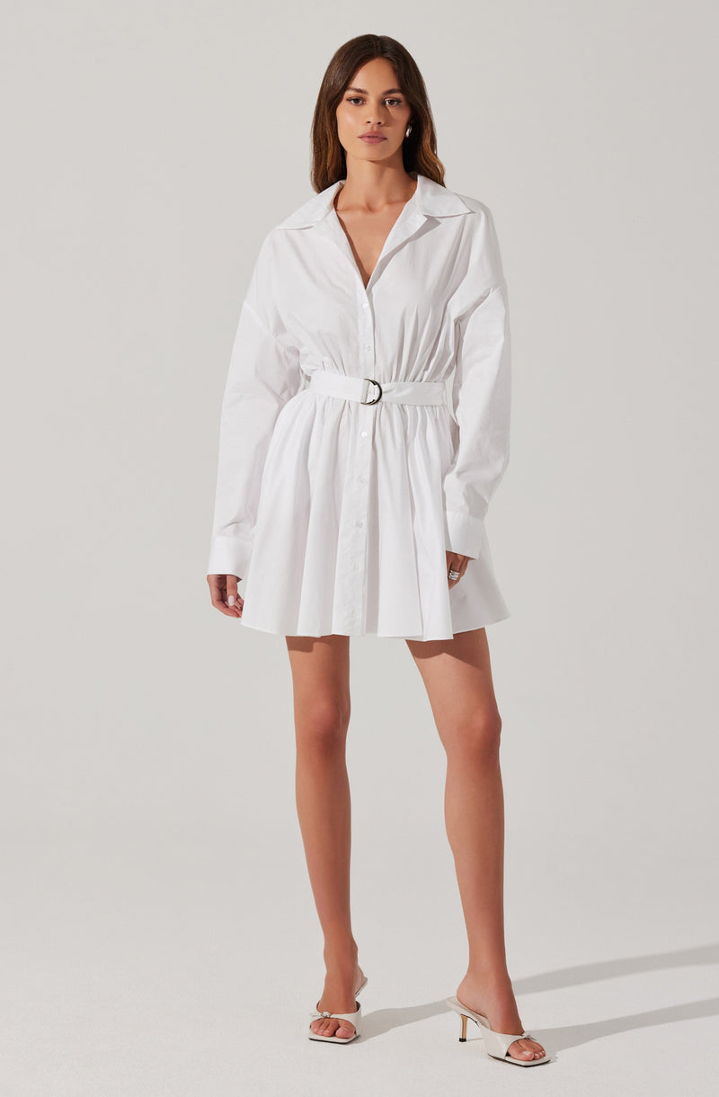A woman is wearing a white shirt dress with a collar, long sleeves, and a waist belt. The dress has a button-front closure and a gathered skirt that falls above the knees. She is also wearing white high-heeled sandals.