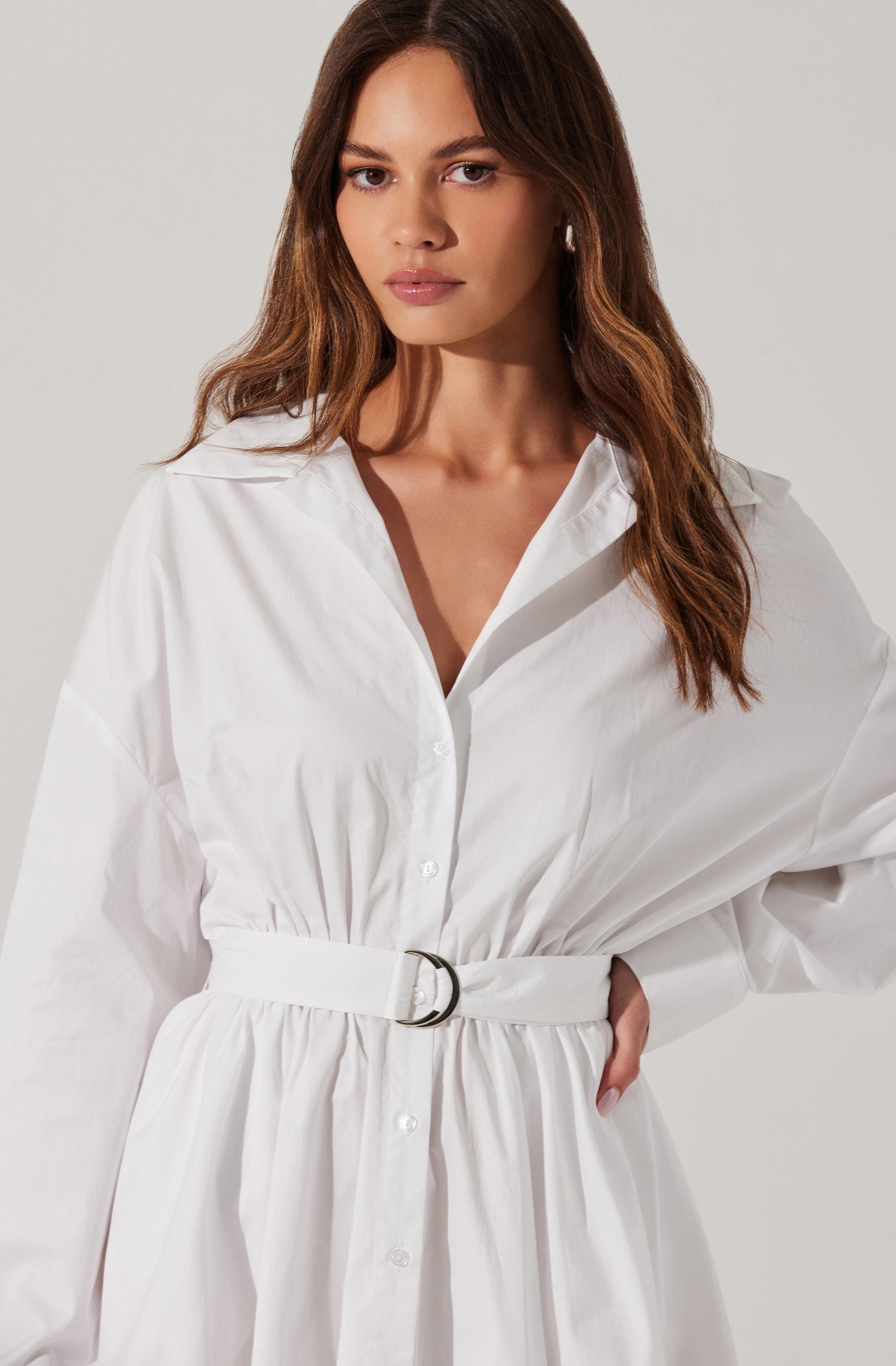 A woman is wearing a white long-sleeve button-up dress with a gathered waist. The dress features a collar and a belt with a silver buckle, creating a cinched silhouette. The fabric appears crisp and slightly textured.