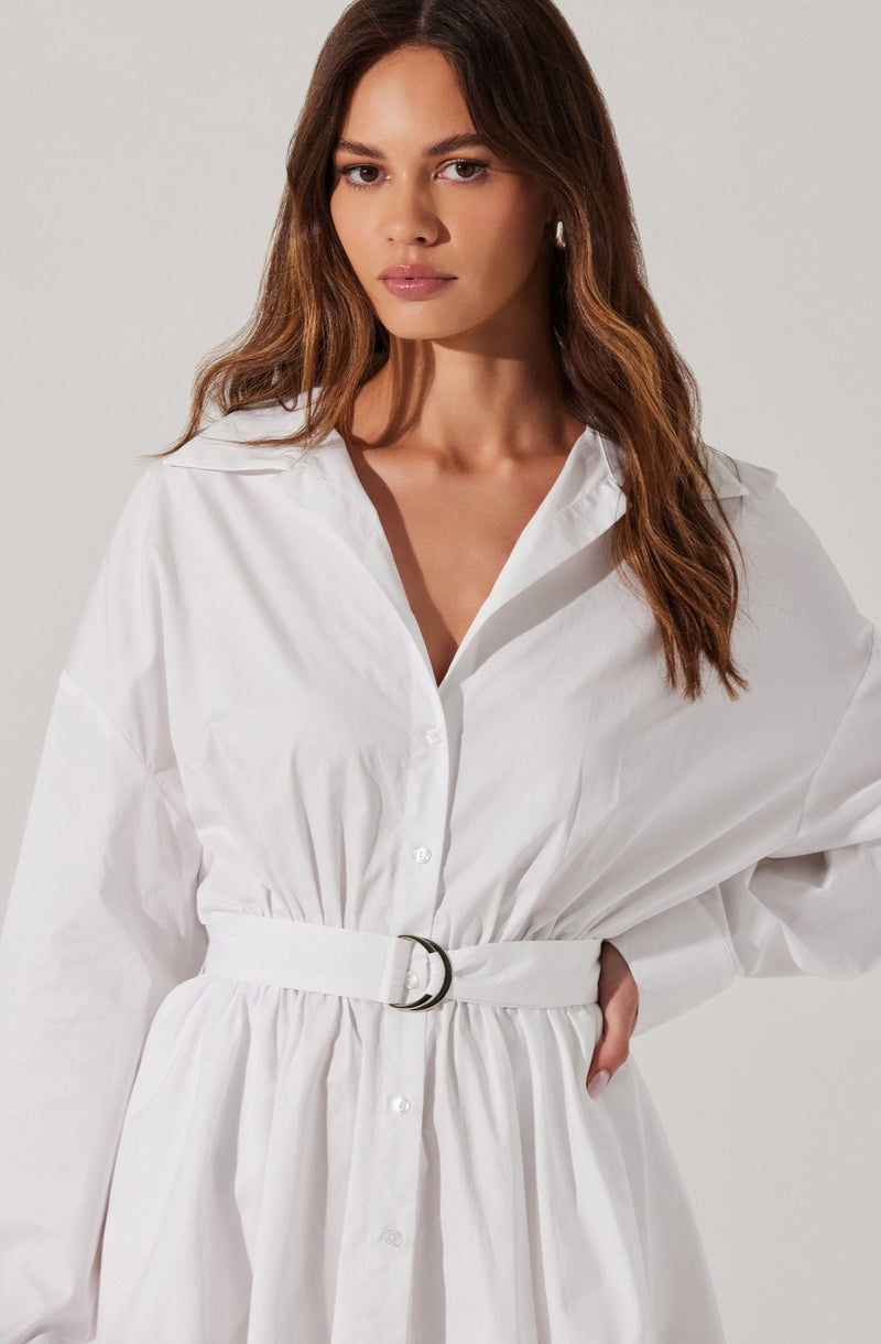 A woman is wearing a white long-sleeve button-up dress with a gathered waist. The dress features a collar and a belt with a silver buckle, creating a cinched silhouette. The fabric appears crisp and slightly textured.