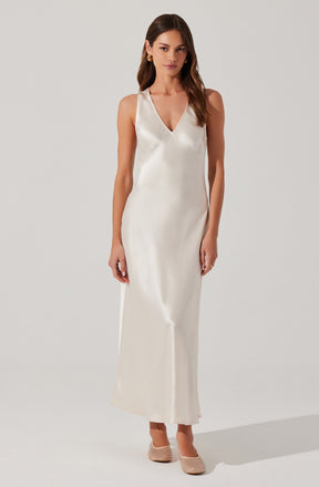 A woman is wearing a sleeveless, full-length, cream-colored satin dress with a V-neckline. She has on gold hoop earrings and nude-colored woven heels.