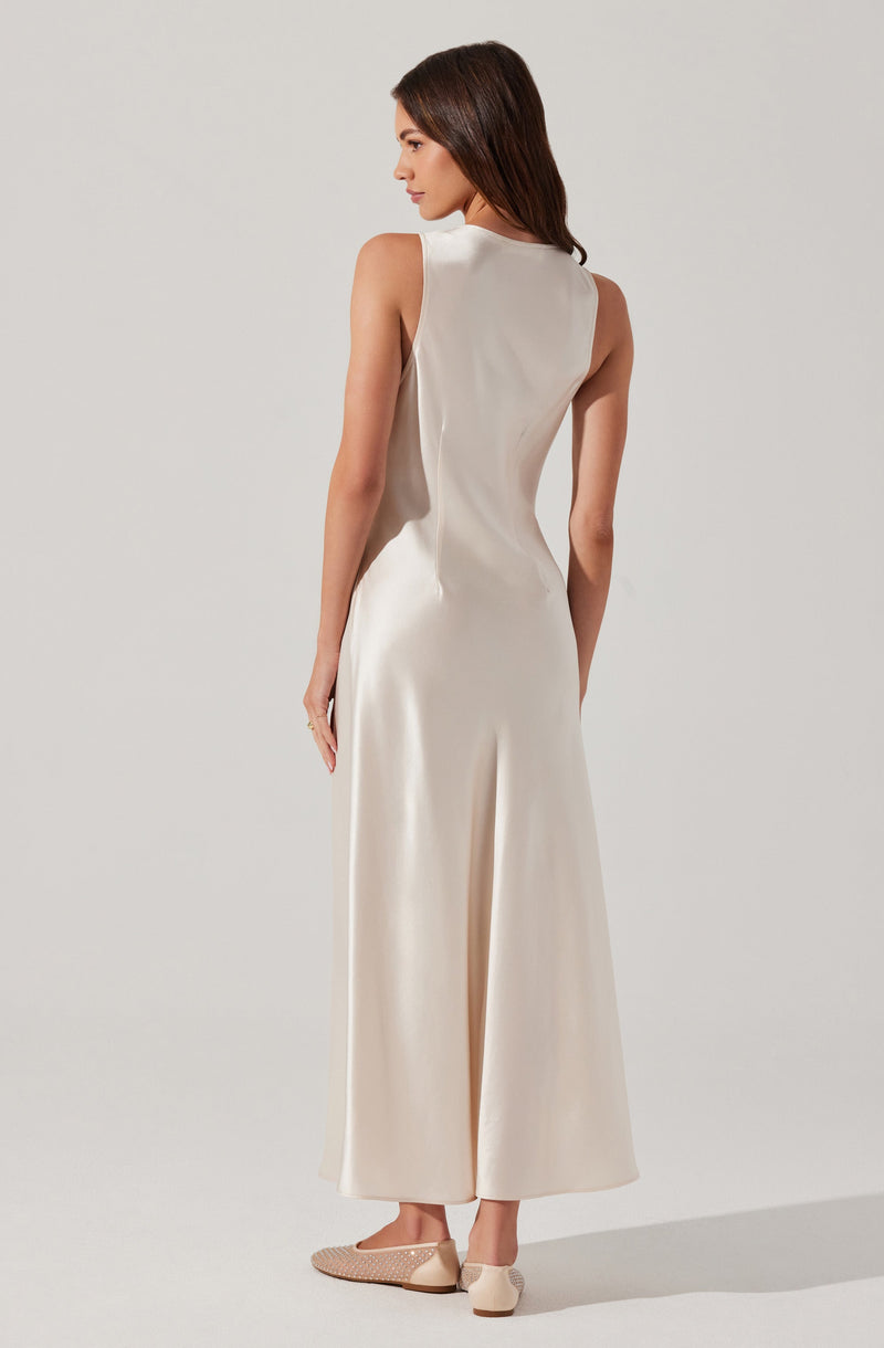 A woman is wearing a sleeveless, high-neck, floor-length cream-colored dress made of a smooth, shiny fabric. She is also wearing beige flats with a textured pattern.