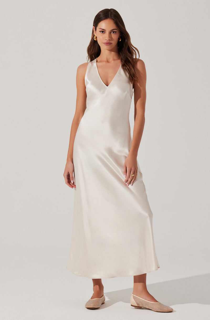 A woman is wearing a sleeveless, V-neck, white satin maxi dress that falls to her ankles. She has paired the dress with gold hoop earrings and beige woven flats.