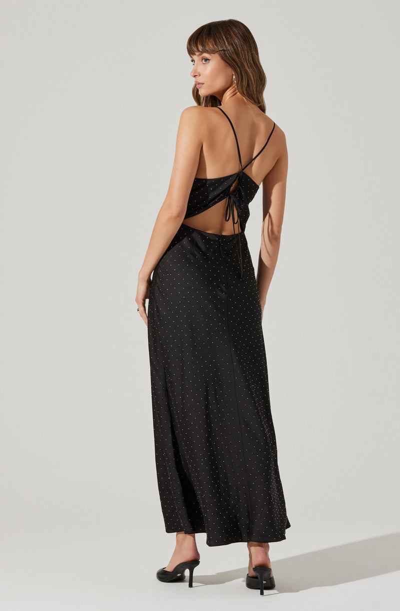 A woman is wearing a black, sleeveless, backless dress with thin spaghetti straps that cross over her shoulders and tie at the lower back. The dress has a subtle polka dot pattern and a floor-length hem. She is also wearing black high-heeled shoes.