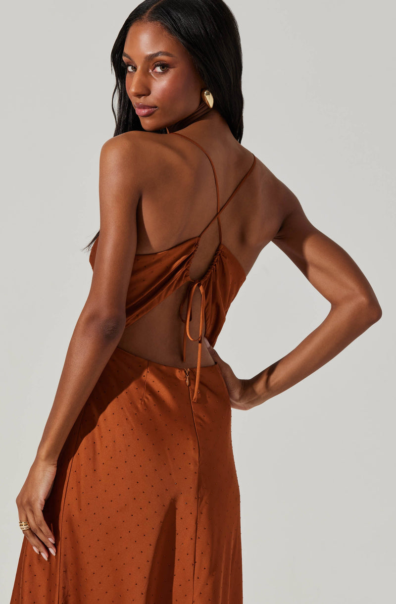 A woman is wearing a silky, rust-colored dress with thin spaghetti straps. The dress features an open back with a gathered detail and a side tie. The fabric appears smooth and shiny, with small perforated dots on the skirt area. She is accessorized with a large, statement earring and a ring on her finger.