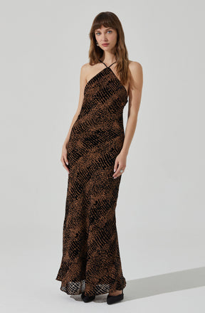 A woman is wearing a full-length, sleeveless dress with a fitted bodice and a flowing skirt. The dress features a black and brown animal print pattern with a mix of small and large scales, creating a textured appearance. The neckline is a halter style with thin straps that cross at the front.